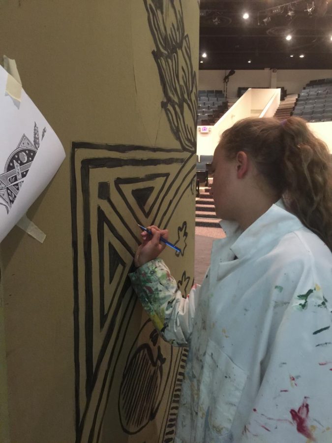 Junior, Kiki Hood, is painting the detailed design on the paper onto the standing cardboard which is a significant part of the set up.